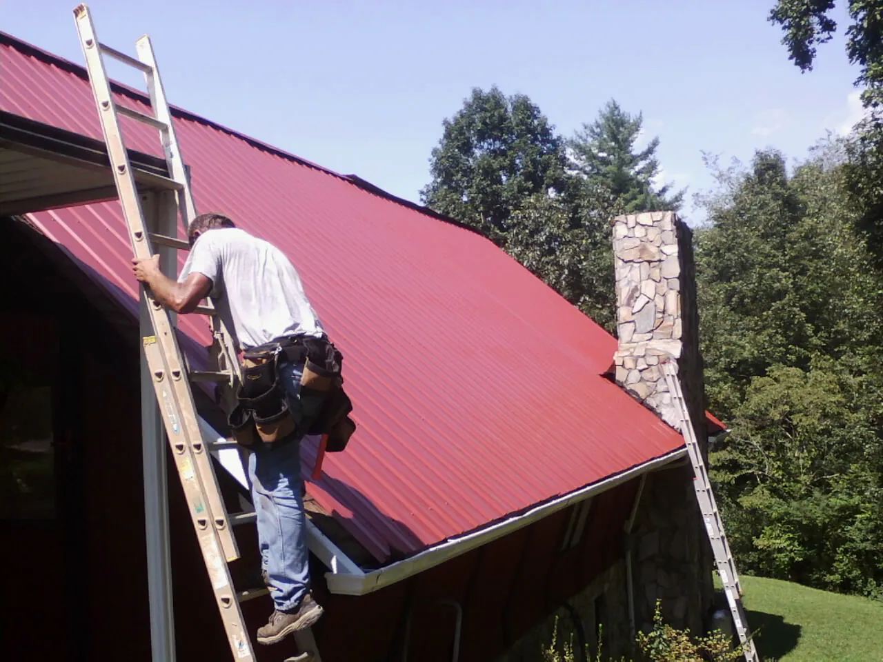 Roofer on a ladder beside a red metal roof and stone chimney — Soffit & Fascia Repair by Roofing Brothers in Vineland